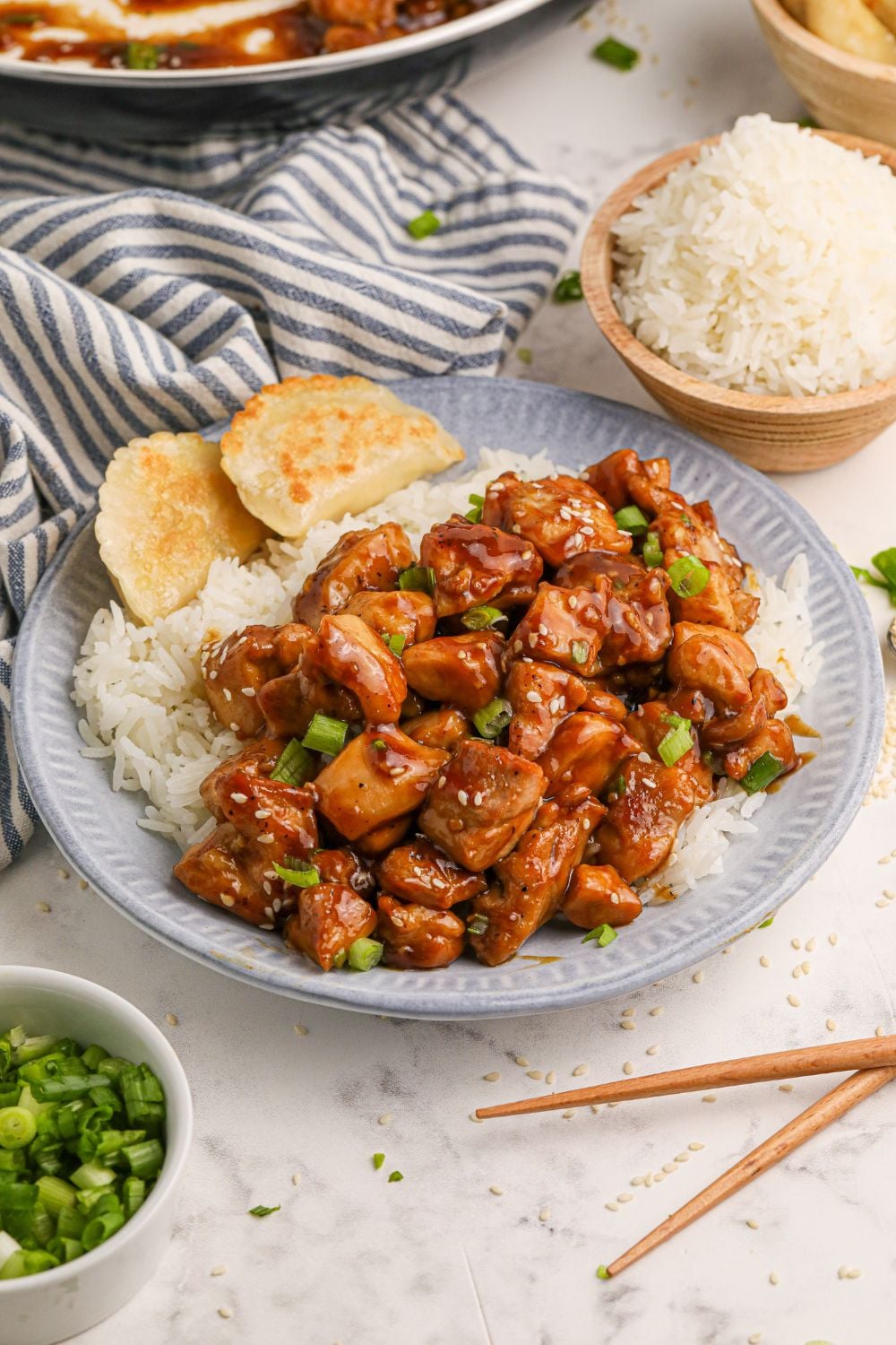 a table full of bourbon chicken on plates, in a skillet, a bowl of green onions to the side, white rice in a bowl, and more