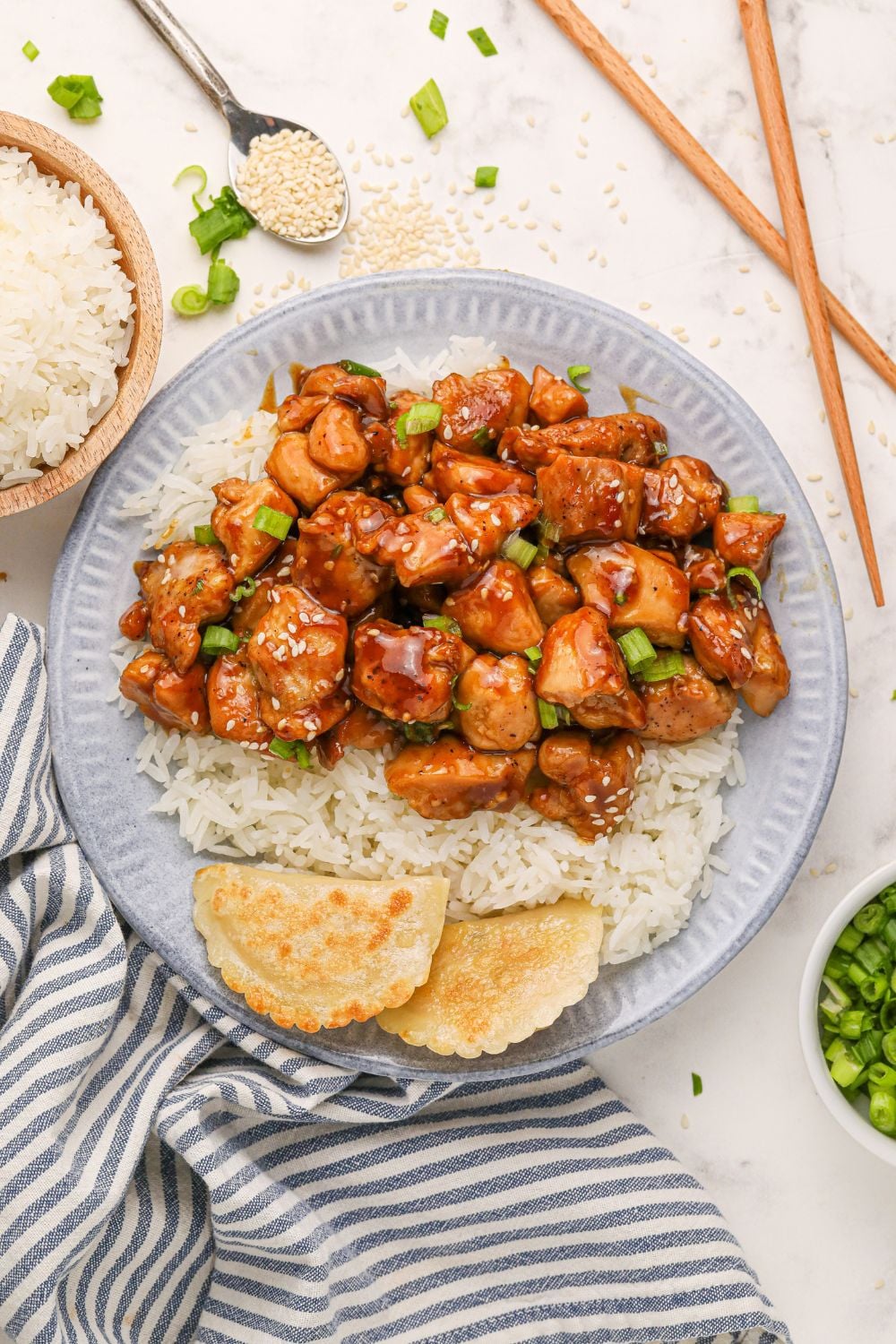 looking down on table with a grey plate with bourbon chicken, rice, and pieorgies to side and a bowl of rice and green onions to side on the table 
