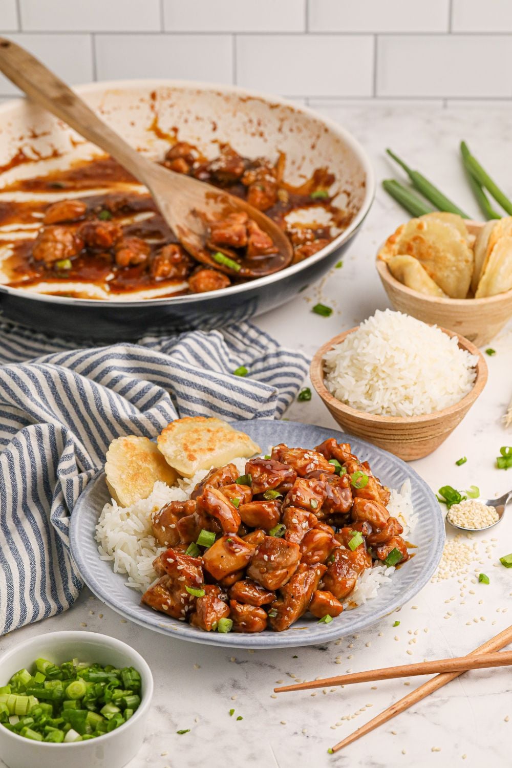 Bourbon Chicken in a skillet with a plate full of chicken and rice in front, a bowl of rice, and pieorgi's as well all on a dinner table 